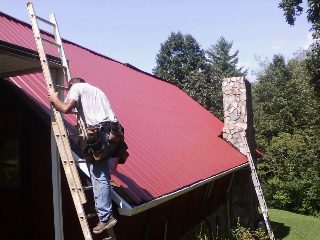 Roofer on a ladder beside a red metal roof and stone chimney — Soffit & Fascia Repair by Roofing Brothers in Marydel